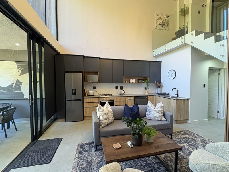 Modern open-plan living area featuring a sleek kitchen with dark cabinetry, integrated appliances, high ceilings, and a glass-railed staircase leading to a mezzanine level.