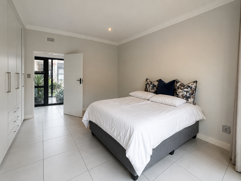 A bright and modern bedroom featuring neutral tiled flooring, extensive built-in cupboards, and direct access to an outdoor area through a glass sliding door.