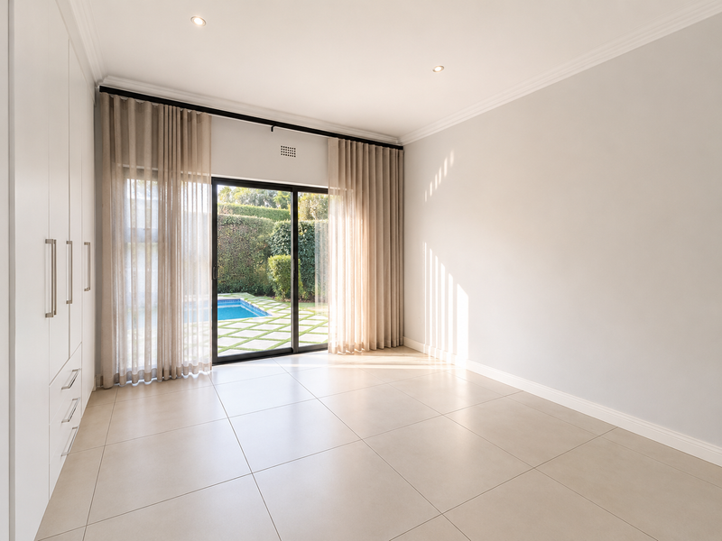 A bright, empty bedroom featuring built-in cupboards, tiled flooring, and a sliding glass door providing direct access to the pool area.