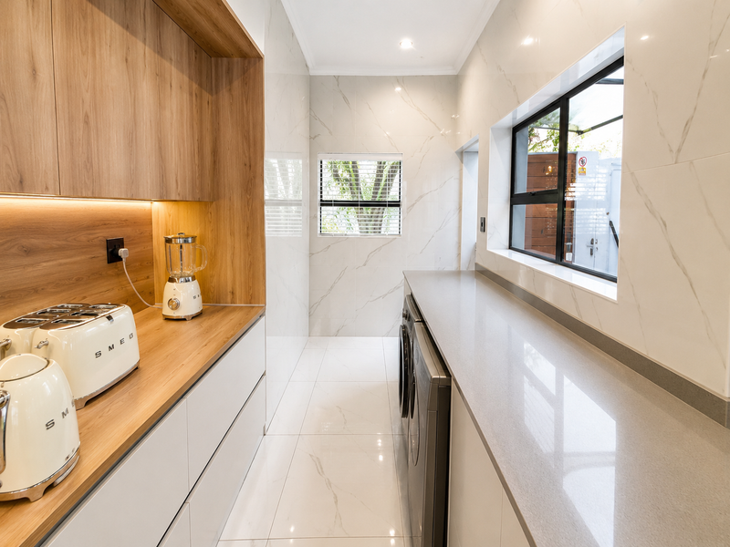 A modern, high-end laundry room featuring sleek built-in cabinetry, stone countertops, marble-look wall tiles, and integrated appliances with excellent natural lighting.