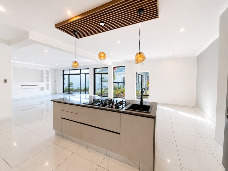 Modern open-plan living area featuring a sleek kitchen island with gas hob, timber slat ceiling detail, and large windows overlooking the garden.