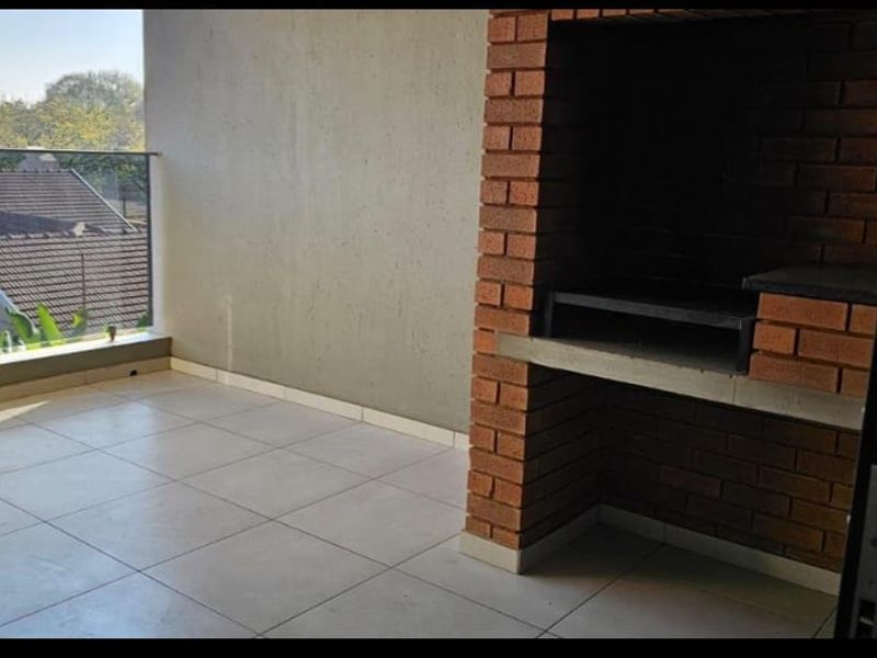 A spacious balcony featuring a built-in brick braai, tiled flooring, and a modern glass balustrade overlooking the surrounding neighborhood.