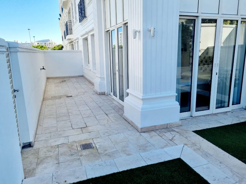 A spacious ground-floor patio featuring light-toned stone tiling, external lighting, and direct access to the apartment interior through large sliding glass doors.