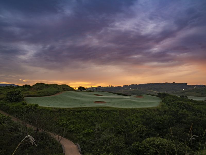 A scenic view of a manicured golf course green set against a dramatic sunset sky, featuring rolling hills and a paved pathway.