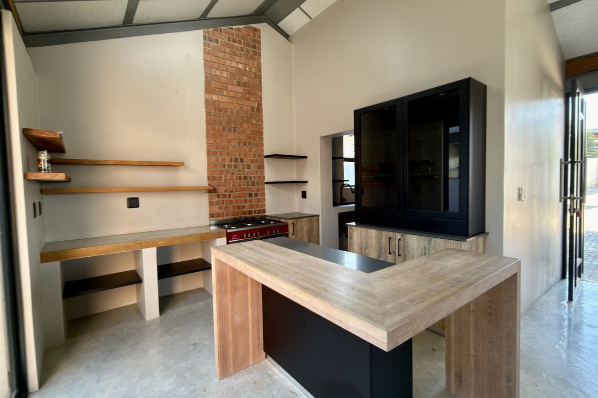 Modern kitchen featuring a striking exposed brick chimney breast, gas stove, timber shelving, and a central island with polished concrete floors.