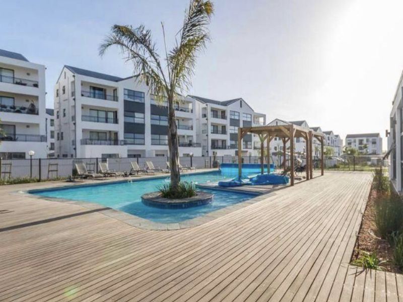 Communal outdoor pool area featuring a swimming pool with a central palm island, surrounded by extensive timber decking and a wooden pergola.
