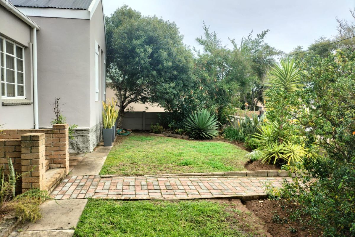 A rear garden area featuring a brick pathway, established landscaping, and a view of the side elevation of the house with a corrugated metal roof.