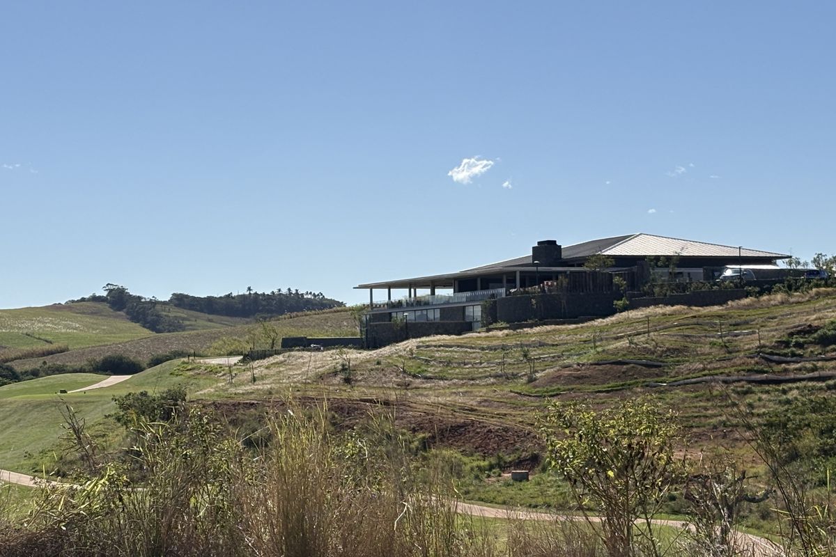 A modern residence featuring stone cladding and a metal roof, situated on a landscaped hillside with a spacious covered patio and glass balustrade.