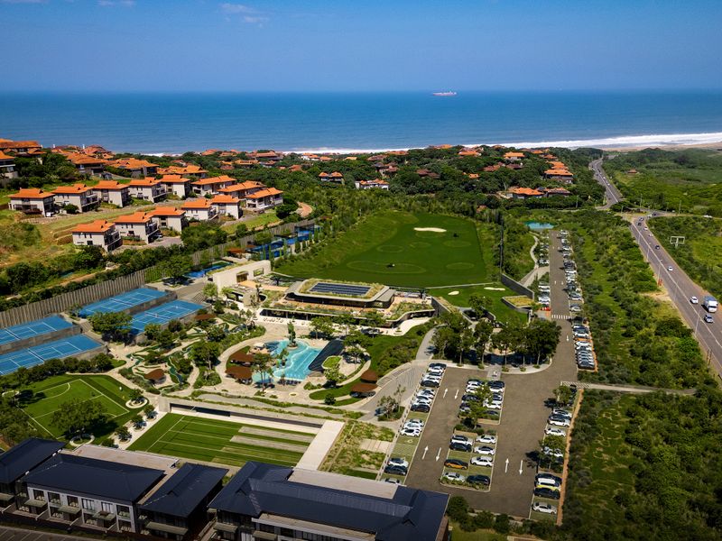 Aerial view of a residential estate featuring a swimming pool, tennis courts, golf course, and extensive parking facilities near the coastline.