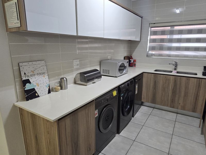 Modern kitchen featuring wood-grain cabinetry, white stone countertops, tiled backsplash, double sink, and integrated laundry appliances.