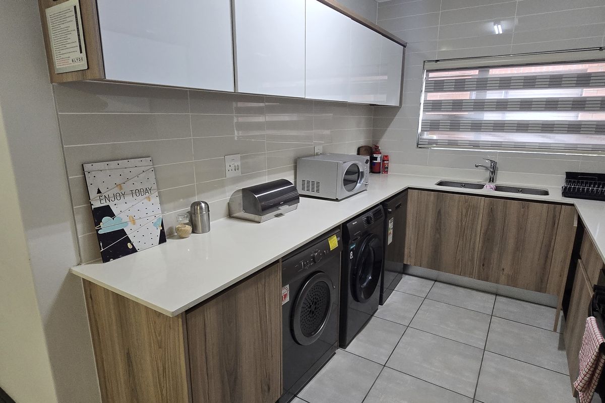 Modern kitchen featuring wood-grain cabinetry, white stone countertops, tiled backsplash, double sink, and integrated laundry appliances.