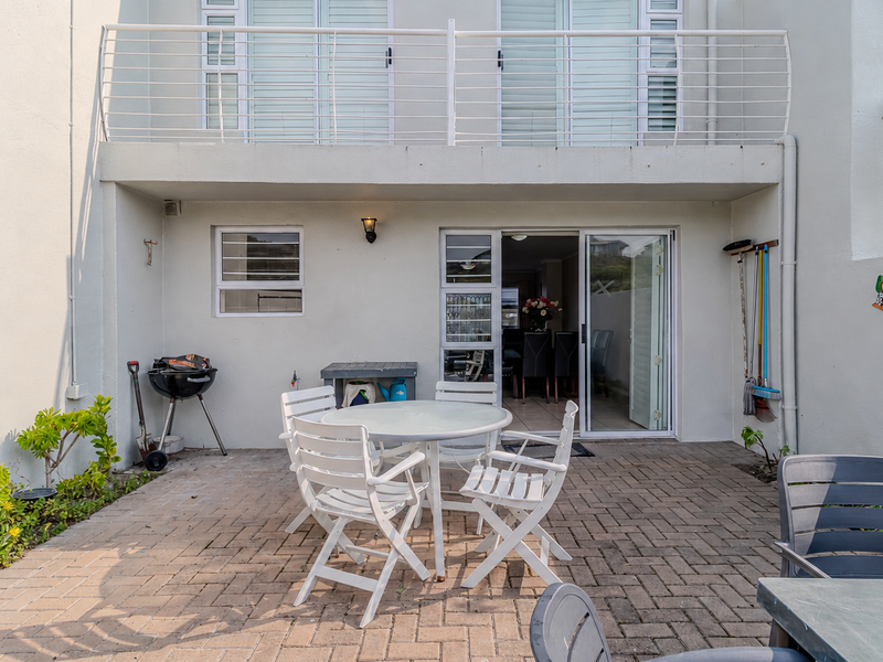 A paved rear patio area featuring a sliding door entrance, an upper balcony with metal railings, and exterior wall lighting.