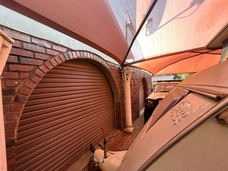 A covered outdoor parking area featuring brick archways, roller shutter garage doors, and a shade sail structure over paved ground.