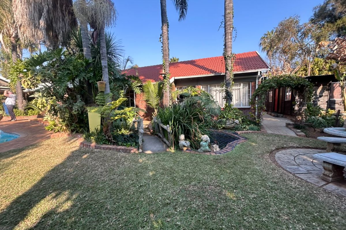 A rear garden view featuring a brick house with a red tile roof, a small decorative bridge, landscaped greenery, and a swimming pool.