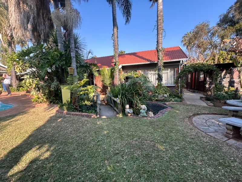A rear garden view featuring a brick house with a red tile roof, a small decorative bridge, landscaped greenery, and a swimming pool.