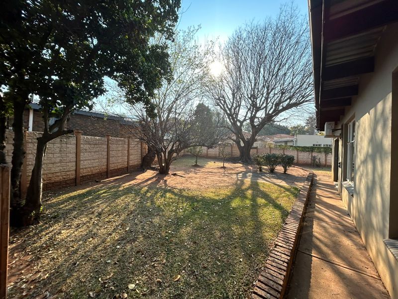 A spacious rear garden featuring a paved walkway, brick edging, and established trees, enclosed by a precast concrete boundary wall.