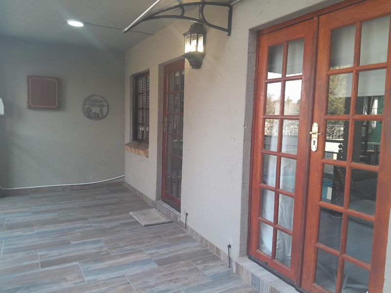 A covered patio area featuring wood-look tiled flooring, timber-framed glass doors, and wall-mounted exterior lighting.