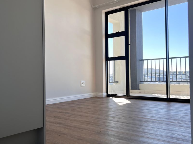 A bright, empty bedroom featuring modern laminate flooring, neutral walls, and a large sliding glass door providing access to a private balcony.