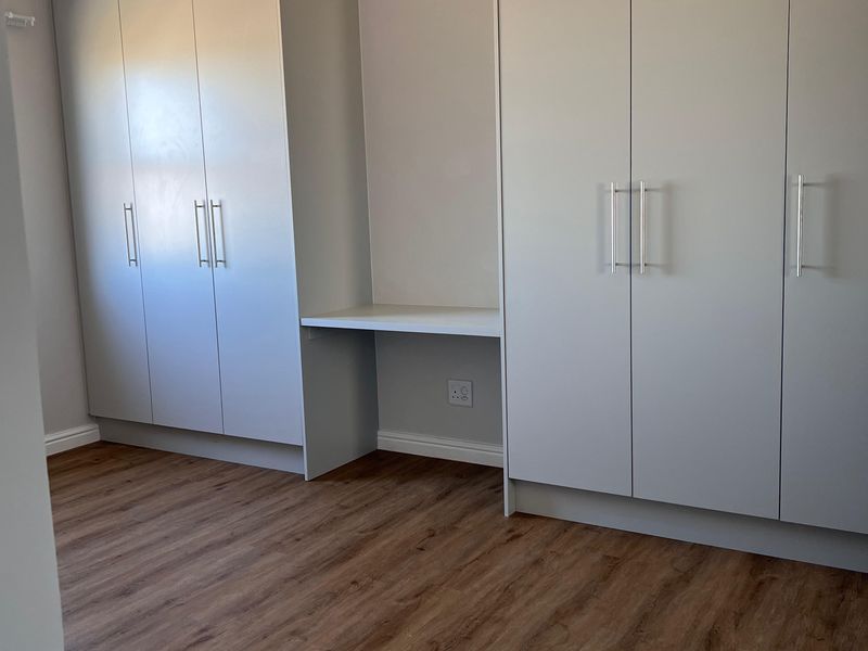 A bright, empty bedroom featuring modern built-in cupboards, a integrated desk area, and wood-look laminate flooring.