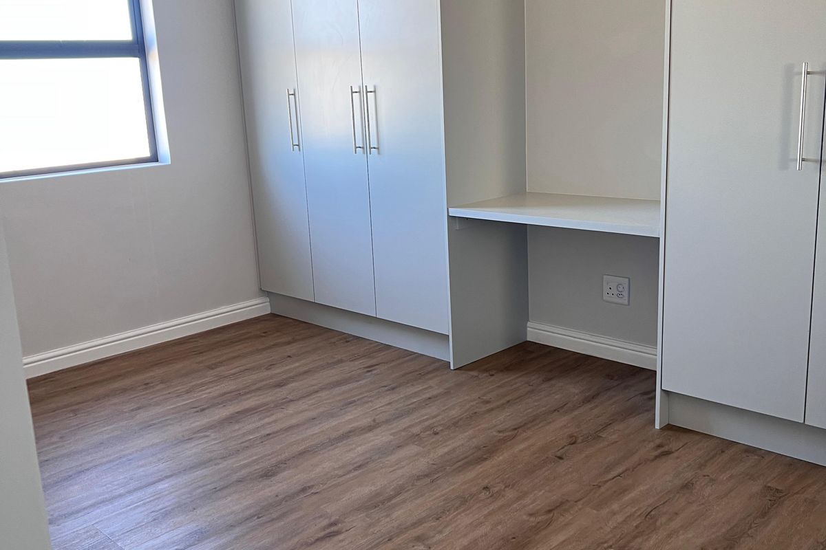 A bright, empty bedroom featuring modern laminate flooring, neutral walls, and extensive built-in cabinetry with an integrated desk space.