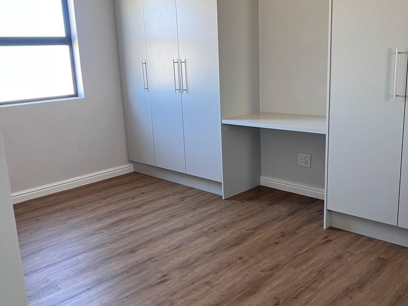 A bright, empty bedroom featuring modern laminate flooring, neutral walls, and extensive built-in cabinetry with an integrated desk space.