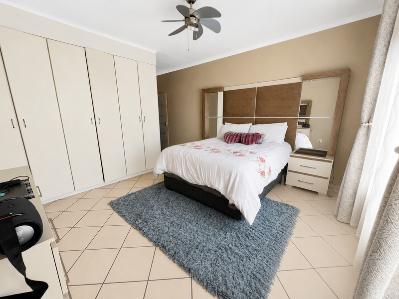 A well-maintained bedroom featuring neutral tiled flooring, extensive built-in cupboard storage, and a modern integrated headboard with a ceiling fan.