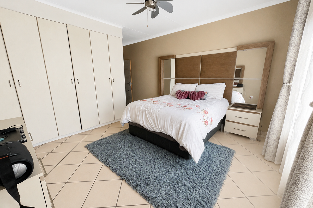 A well-maintained bedroom featuring neutral tiled flooring, extensive built-in cupboard storage, and a modern integrated headboard with a ceiling fan.