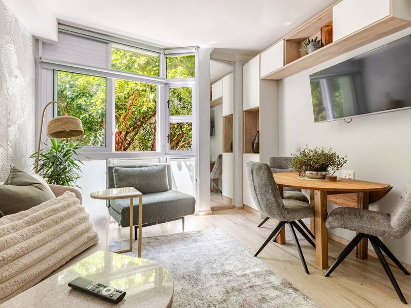 A bright open-plan living and dining area featuring modern built-in cabinetry, light laminate flooring, and large windows overlooking lush greenery.