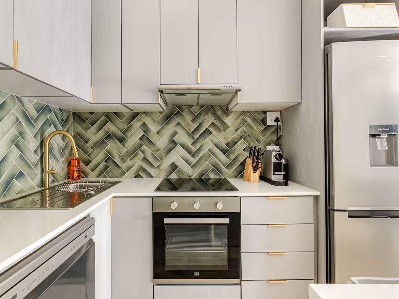 Modern kitchen featuring grey cabinetry, quartz countertops, a stylish green herringbone tile backsplash, and integrated stainless steel appliances.