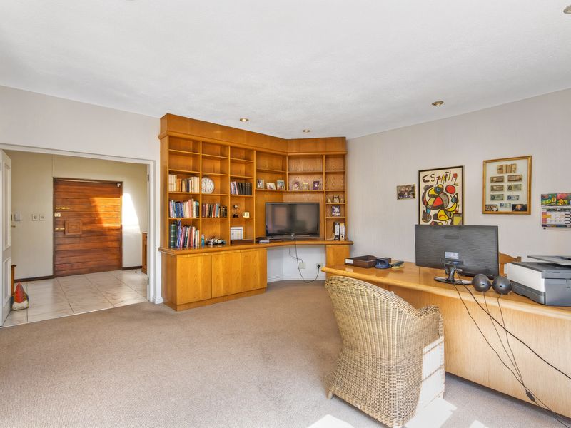 A home office featuring a large built-in wooden shelving unit, recessed lighting, and carpeted flooring, with an adjacent entryway visible.