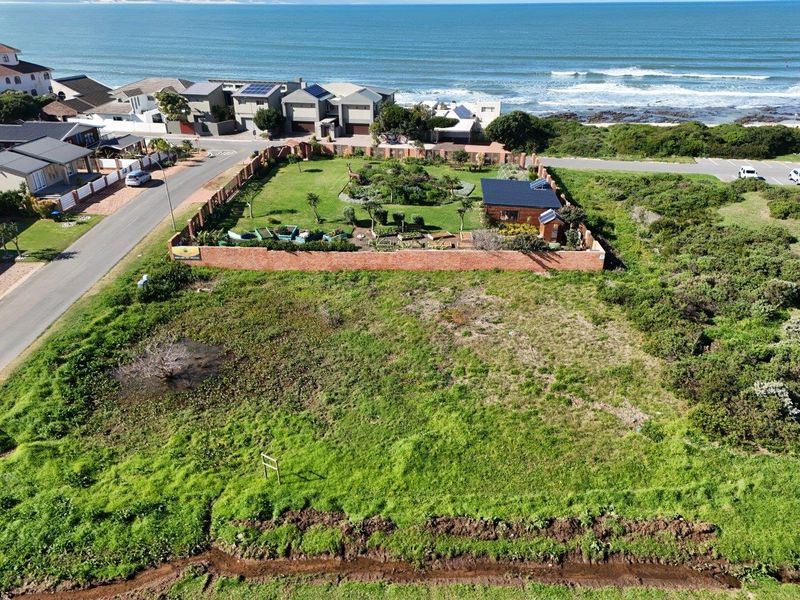 Aerial view of a vacant residential plot featuring a landscaped garden, perimeter brick walling, and a small outbuilding with solar panels near the coast.