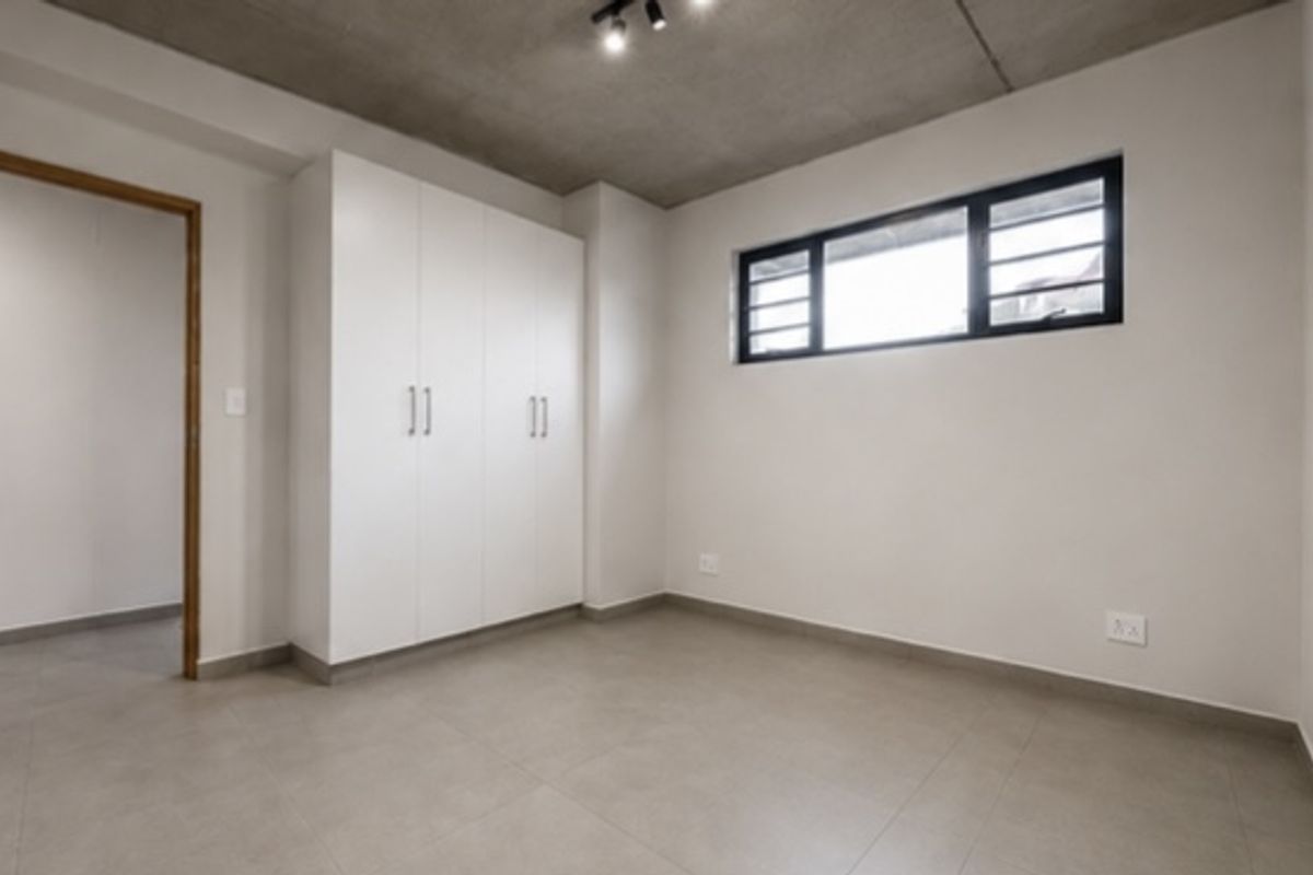 A clean, modern bedroom featuring built-in cupboards, tiled flooring, and an exposed concrete ceiling with track lighting.