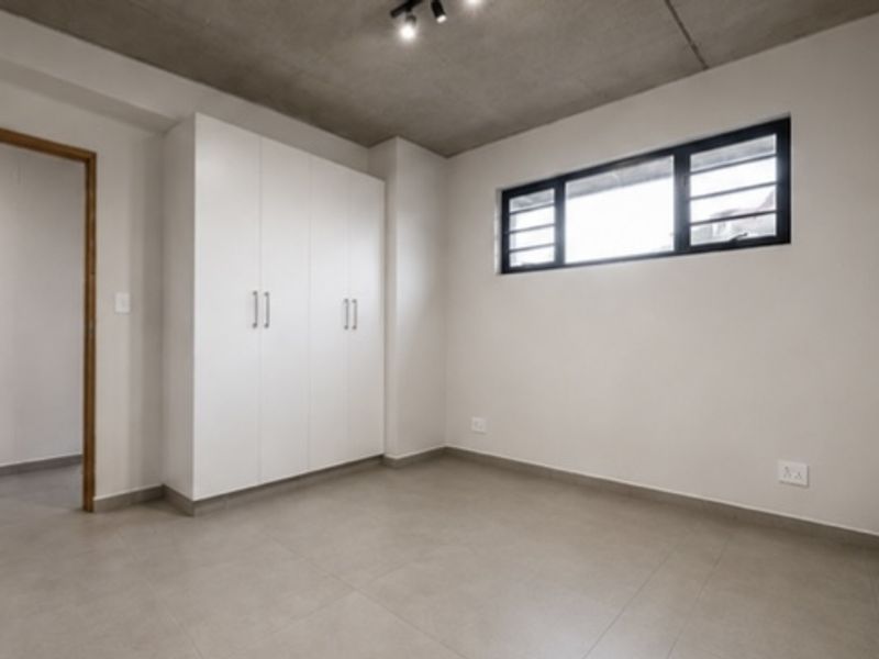 A clean, modern bedroom featuring built-in cupboards, tiled flooring, and an exposed concrete ceiling with track lighting.