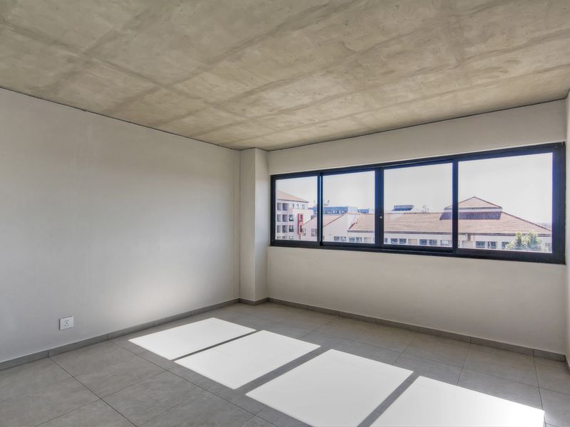 A bright, empty bedroom featuring an exposed concrete ceiling, neutral tiled flooring, and large aluminium-framed windows with urban views.