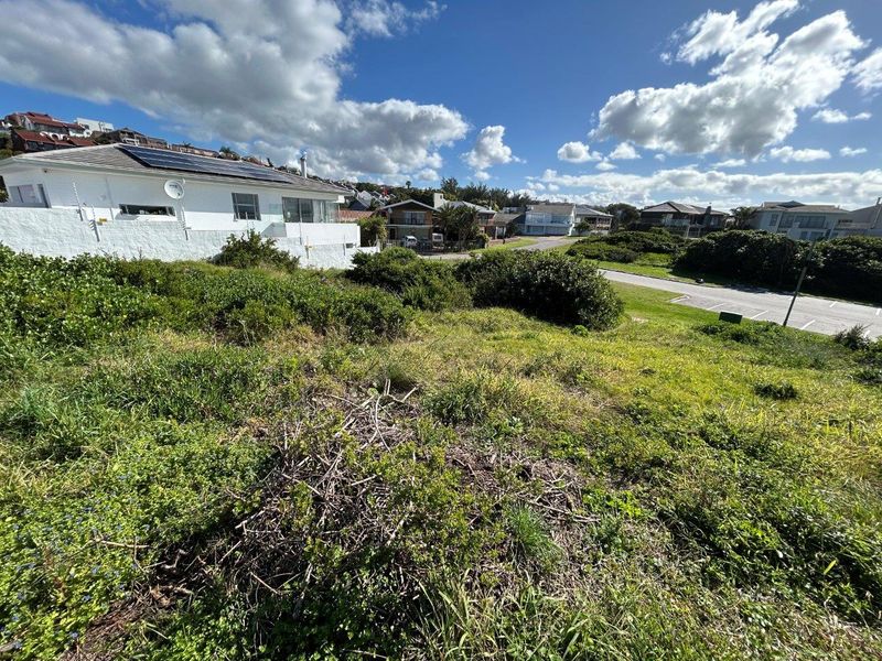 A view of a vacant residential plot featuring sloped terrain and natural vegetation, situated in a developed coastal neighborhood.