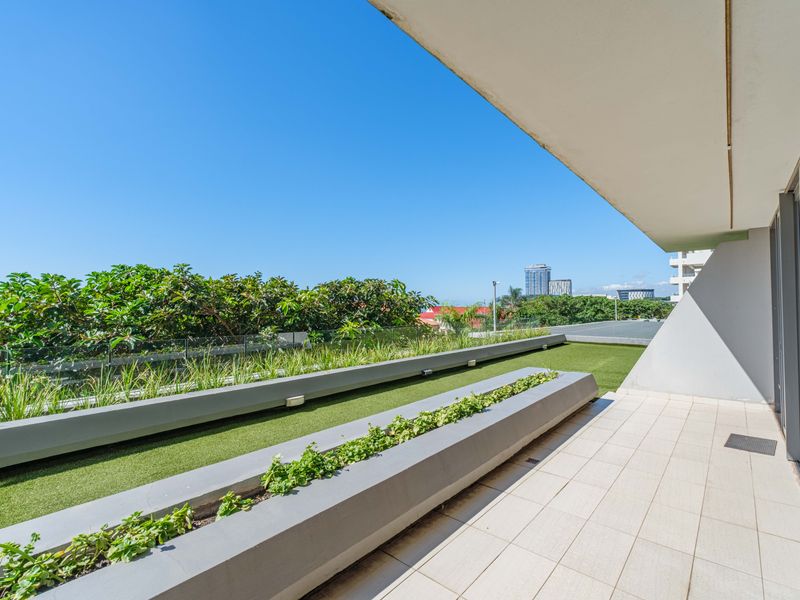 Spacious apartment balcony featuring tiled flooring, built-in concrete planters, artificial turf, and a glass balustrade with clear urban views.