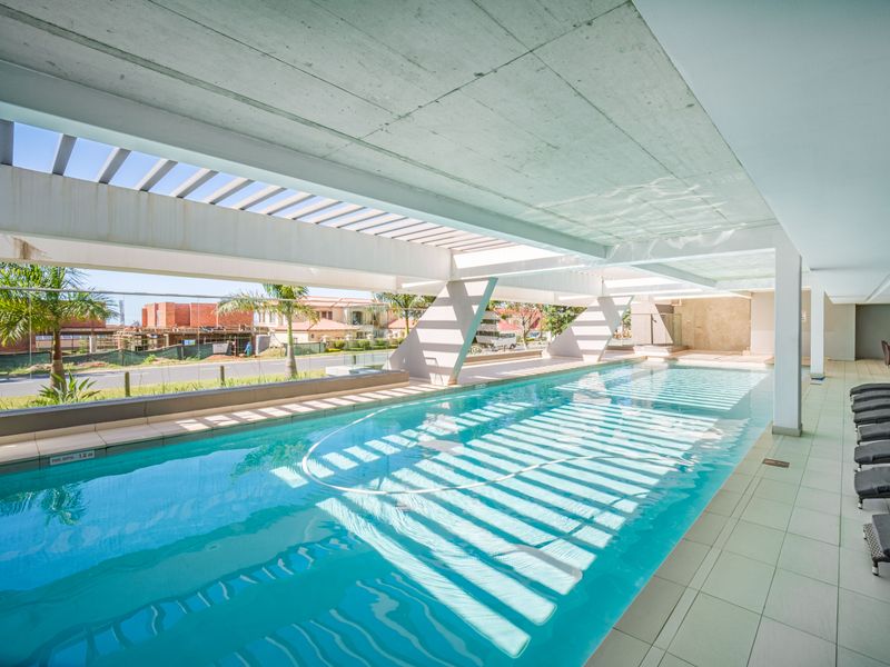 A modern, covered swimming pool area featuring a concrete pergola, tiled decking, and a glass balustrade overlooking the surrounding neighborhood.