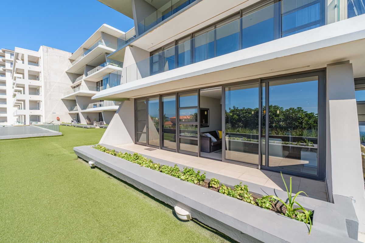 Modern apartment exterior featuring a spacious patio with glass sliding doors, glass balustrades, and a built-in concrete planter box overlooking an artificial lawn area.