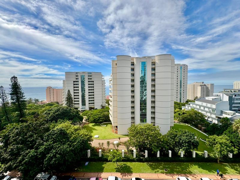 An elevated aerial view of a modern apartment complex featuring glass elevator shafts, surrounded by lush green gardens and coastal scenery.