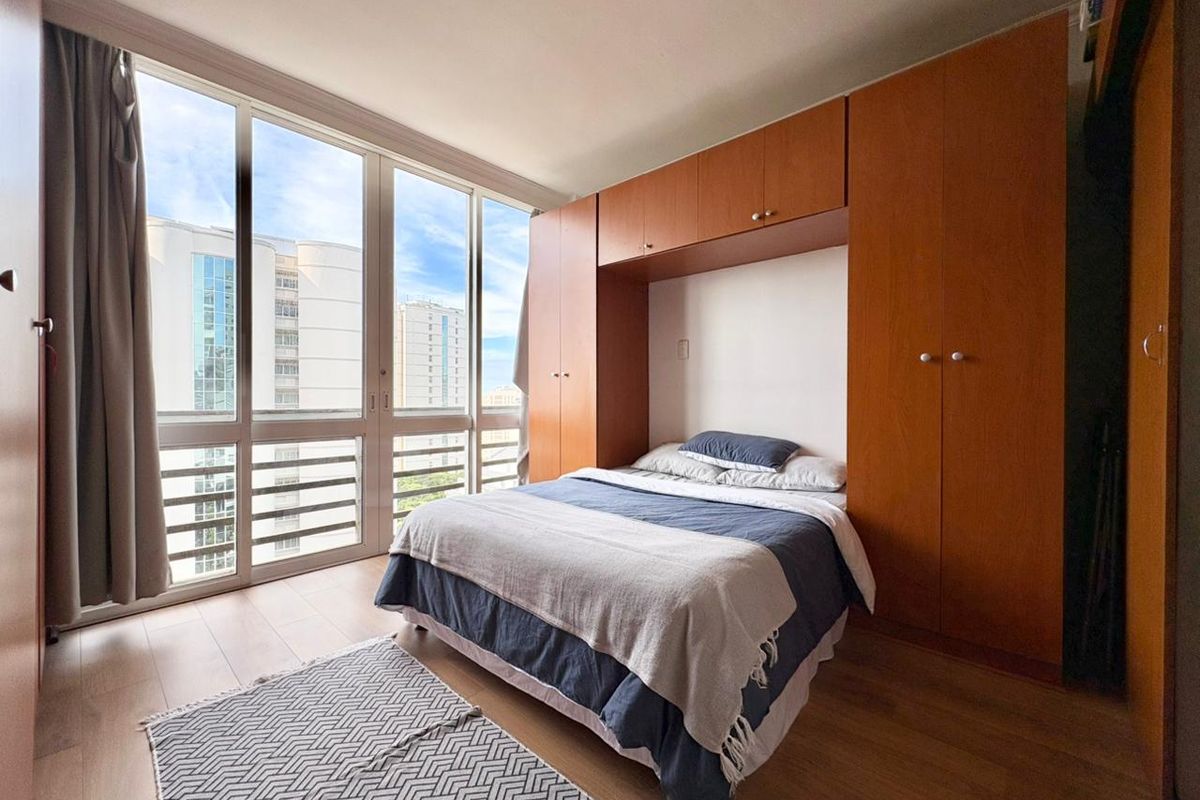 A bright bedroom featuring floor-to-ceiling windows with city views and extensive built-in wooden cabinetry for storage.