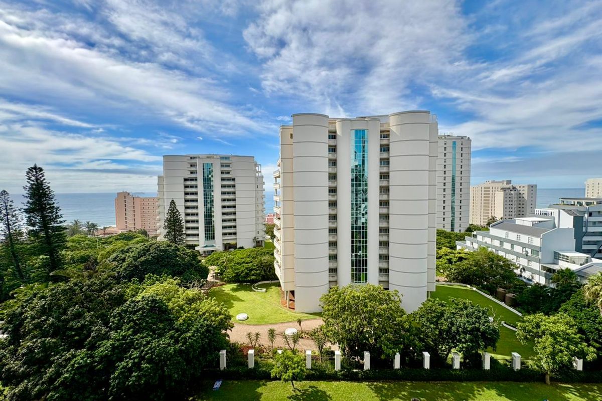 An elevated aerial view of a modern apartment complex featuring glass elevator shafts, surrounded by lush green gardens and coastal scenery.