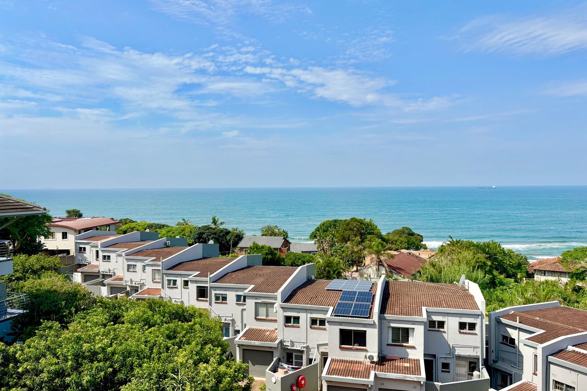 An elevated coastal view overlooking a residential complex with tiled roofs and solar panels, set against a clear blue ocean horizon.
