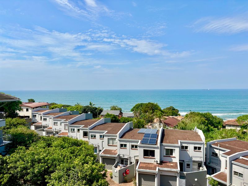 An elevated coastal view overlooking a residential complex with tiled roofs and solar panels, set against a clear blue ocean horizon.