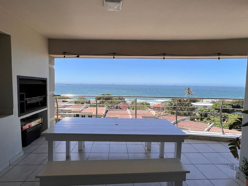 Spacious covered balcony featuring a built-in braai, tiled flooring, and a stainless steel balustrade, offering expansive ocean and coastal views.
