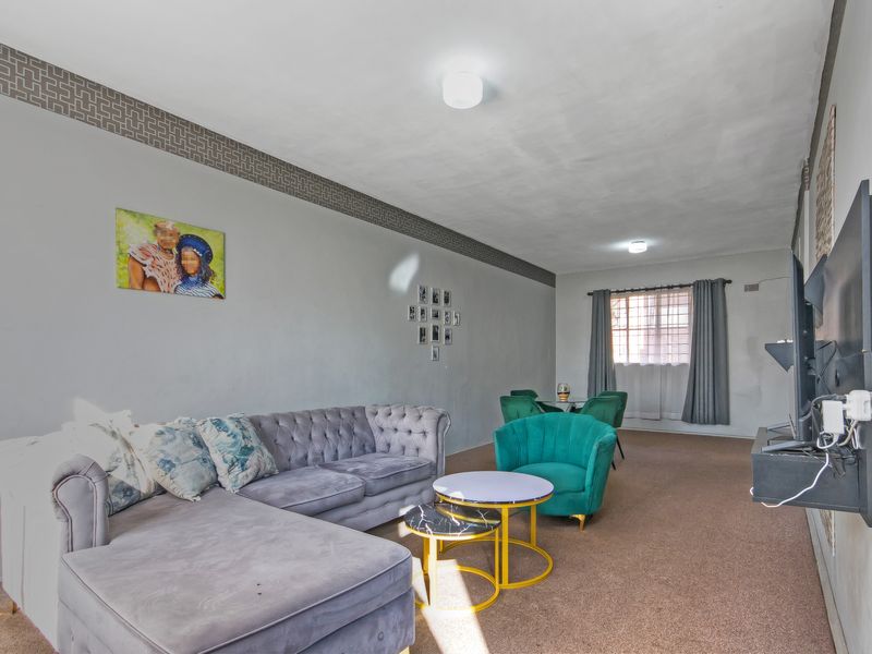 A spacious living room featuring neutral walls with decorative border detailing, carpeted flooring, and a window fitted with security bars.