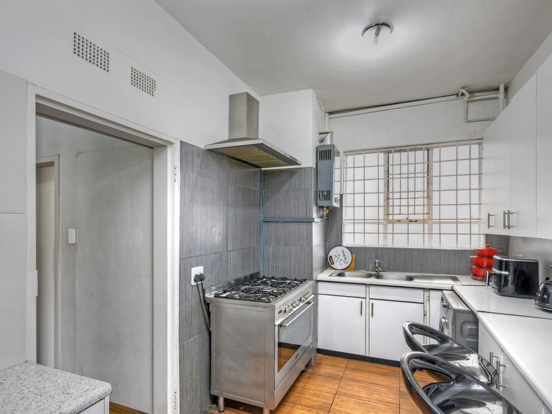 A functional kitchen featuring a gas stove, extractor fan, stainless steel sink, and built-in cabinetry with a tiled splashback and security window bars.
