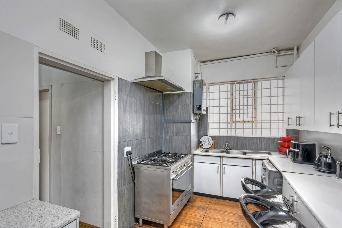 A functional kitchen featuring a gas stove, extractor fan, stainless steel sink, and built-in cabinetry with a tiled splashback and security window bars.