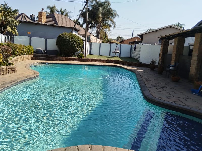 A well-maintained swimming pool area featuring a paved deck, brick retaining walls, and an adjacent covered patio space within a secure walled garden.