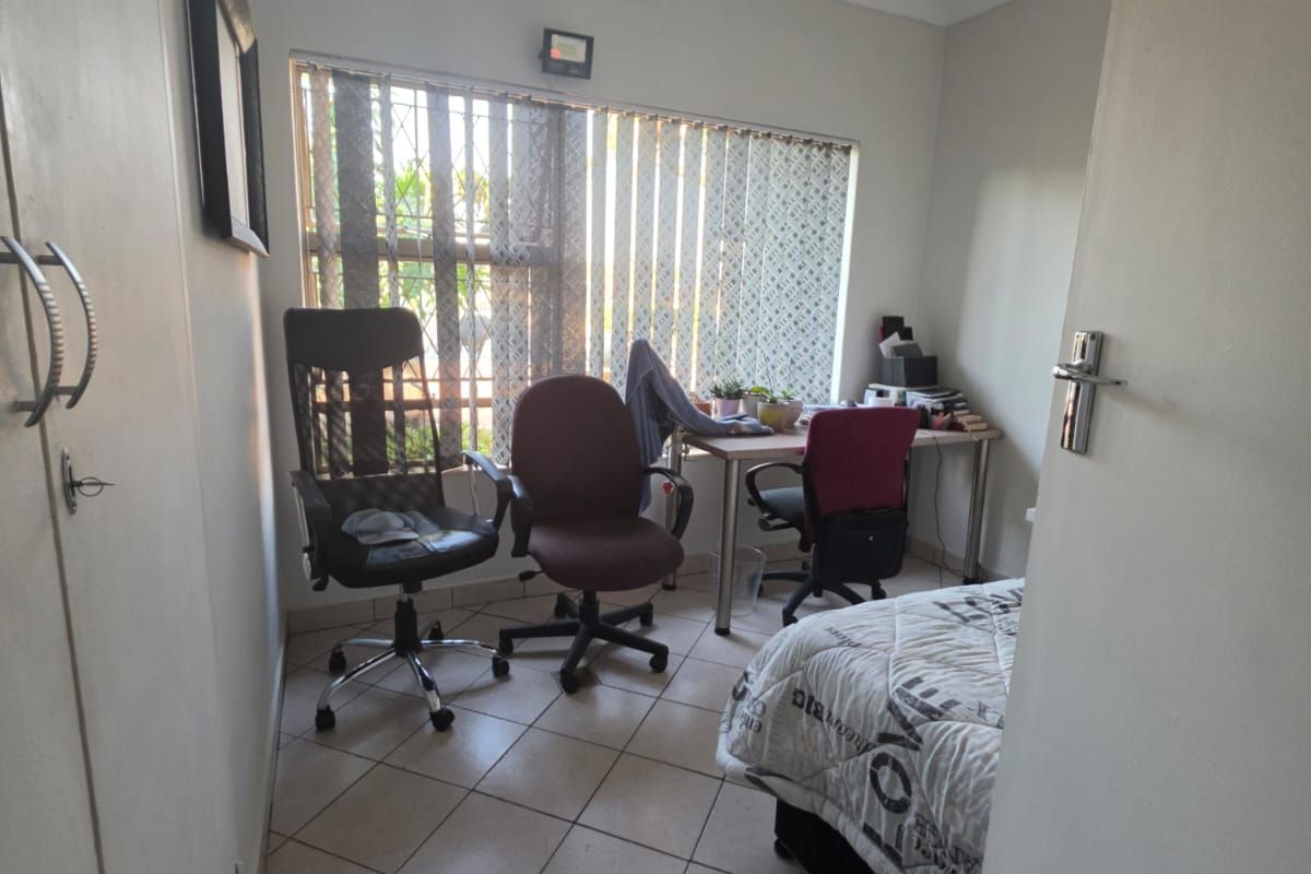 A compact bedroom featuring tiled flooring, a built-in cupboard, and a window with vertical blinds and security bars.