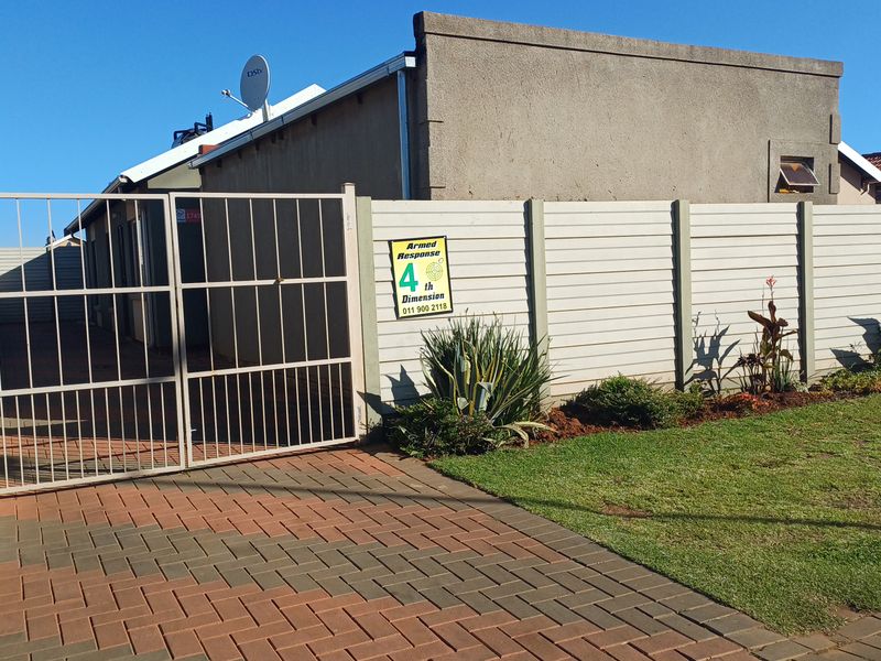 Front exterior view of the property featuring a paved driveway, metal security gate, and precast concrete boundary wall with established garden landscaping.